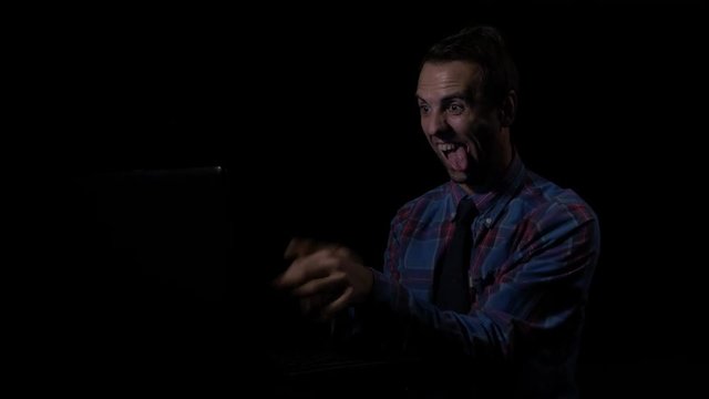 Happy smiling male hacker doing funny crazy gestures with his tongue and face after cracking codes in a dark room