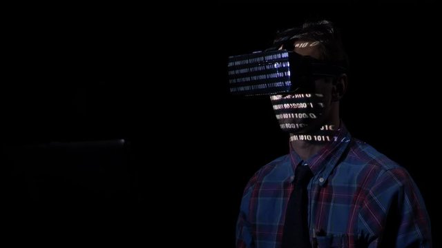 Software developer experiencing virtual reality goggle glasses at night in a dark room to read binary codes reflected on his face