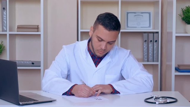 doctor sitting at the desk writing prescription for some medicine. mixed race therapist using laptop checking information
