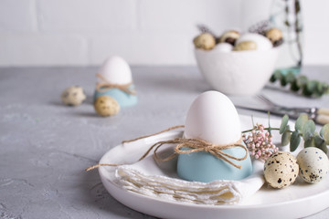 Fototapeta premium Easter festive table setting with white chicken eggs in eggs cups, leaf sprigs of eucalyptus. On a gray concrete background.