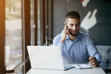 Naklejka premium Young handsome man sitting in office with cup of coffee and working on project connected with modern cyber technologies. Businessman with notebook trying to keep deadline in digital marketing sphere.