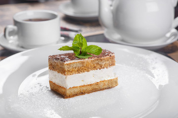 Rectangular piece of Tiramisu with a sprig of mint on a white plate. In a low key, in the background a cup of tea and a kettle, cutlery. Serving, serving dishes in a restaurant, cafe, bar
