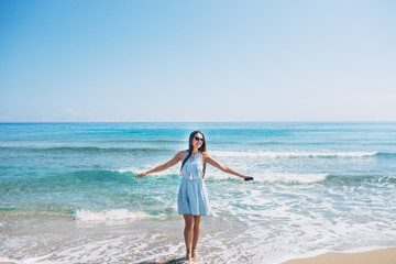 Smiling girl with long brown hair and in sunglass dancing on the beach with earphones and phone in hand. Aqua blue sea on background