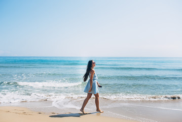 Smiling girl with long brown hair and in sunglass walking barefoot on the beach with earphones and phone in hand. Sea on background