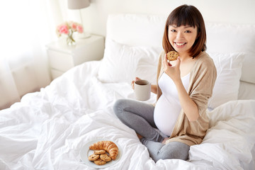 happy pregnant woman eating cookie in bed at home