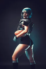 Fotobehang Sportive serious woman in helmet of rugby player holding ball in stuio on dark background. Back view. © zamuruev