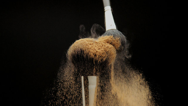 closeup of black brush with a facial powder on it and another brush whisk away a part of powder