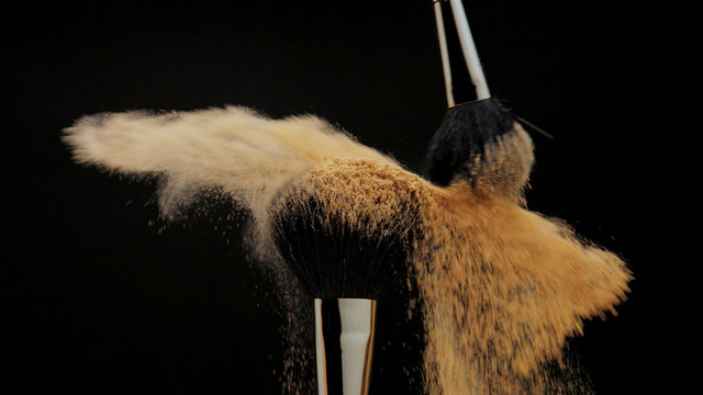 closeup of black brush with a facial powder on it and another brush whisk away a part of powder