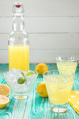 lemonade or limoncello in glasses with ice cubes, sherbet glass with ice cubes, bottle with drink, lemon fruits on turquoise colored wooden table