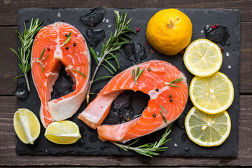 Two fresh raw salmon steaks with salt, peppers, lemon and rosemary with ice cubes on black slate board