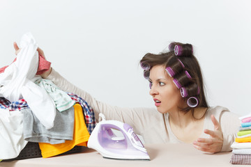 Distressed housewife with curlers on hair in light clothes ironing family clothing on ironing board...