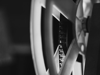 Front view of an old-fashioned antique Super 8mm film projector, projecting a beam of light in a dark room next to a stack of unraveled film reels. Black and white