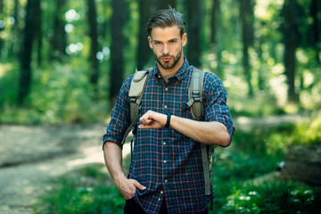 Tourism, travel, adventure, hike concept - handsome bearded man hiking in forest, dressed in checkered shirt looks at clock. Man with beard and moustache traveler with backpack walking in forest.