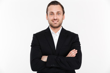 Portrait of successful businessman having stubble standing in suit with broad smile keeping arms folded, isolated over white background