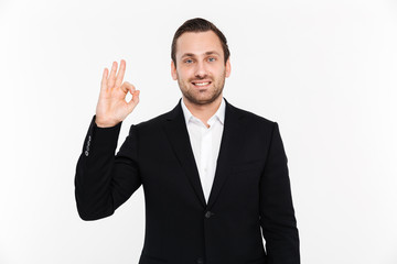 Portrait of smiling entrepreneur having stubble wearing black suit and showing OK sign meaning alright, isolated over white background