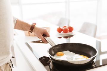Cropped photo of caucasian bachelor in white shirt cooking omelet with vegetables in home kitchen, using frying pan