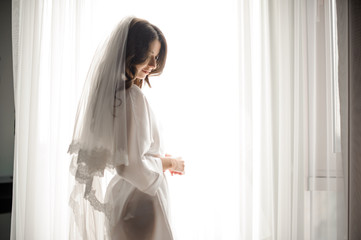 Bride morning preparation. Bride in white wedding negligee and veil near the window