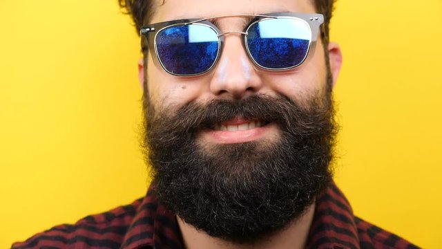 Natural smiling long bearded hipster with his sunglasses on over yellow background in close up