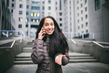 young girl talking on mobile phone in courtyard business center. girl with long dark hair dressed in winter jacket in cold weather speaks on phone on background buildings made of glass and concrete