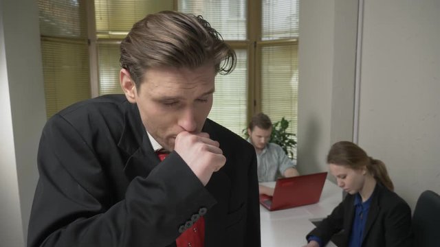 Young man in a suit is coughing, sick, a disease concept. Man and the girl in the background are working in the office. 60 fps