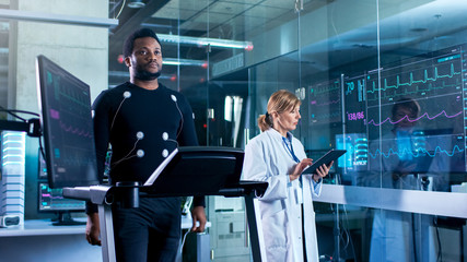 Male Athlete Walks on a Treadmill with Electrodes Attached to His Body while Sport Scientist Supervises Reads EKG Status. In the Background Laboratory with High-Tech Equipment.