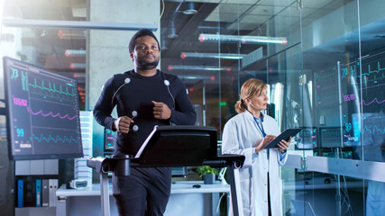 Male Athlete Walks on a Treadmill with Electrodes Attached to His Body while Sport Scientist Interacts with Touchscreen and Supervises EKG Status.