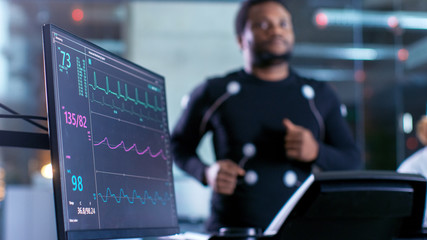 Close-up Shot of a Monitor With EKG Data. Male Athlete Runs on a Treadmill with Electrodes Attached...