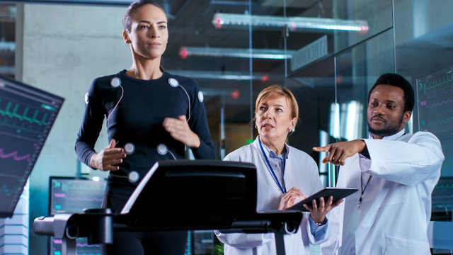 Beautiful Woman Athlete Runs on a Treadmill with Electrodes Attached to Her Body, while Two Scientists Supervise Watching EKG Data Showing on Laboratory Monitors.