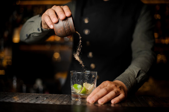 Bartender adding cane sugar into the cocktail glass with lime. Process of making Caipirinha cocktail