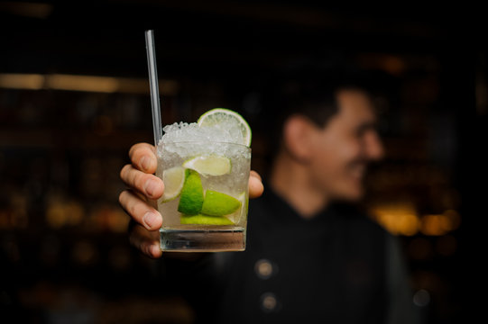 Bartender hand holding a glass filled with Caipirinha cocktail with a straw. Close up