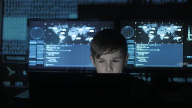 Cute teen boy programmer working at a computer in the data center filled with display screens. Portrait of Child prodigy hacker.