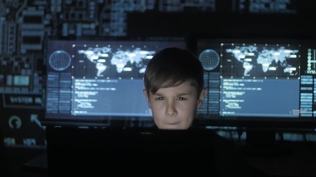 young prodigy boy hacker programmer working at the computer in the data center filled with display screens. Portrait of Child prodigy hacker.