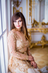 Fototapeta premium Portrait of cute girl in gold dress, sits on window sill, in luxurious room