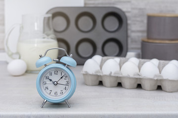 Light big kitchen. Tableware for cooking, cake mold and tray with eggs on the table. Blue alarm clock on table.