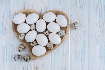 White eggs and quail eggs in wooden heart shaped plate on wooden white background