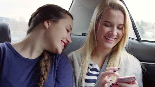 Happy Blonde Women Shows Her Friend Something Funny On Her Smartphone, In Back Seat Of Moving Car - Shot On Red Scarlet-W Dragon In 4K/Slow Motion