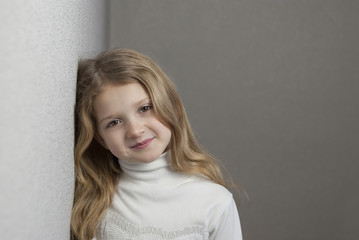Pretty little blonde girl looking in camera and smiling while standing near wall