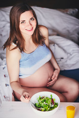 Pregnant smiling girl in T-shirt sit on bed and eat salad with green and red leaves, drink orange, pumpkin and carrot juice. Young woman keep diet during pregnancy. Healthy food for expectant mothers.