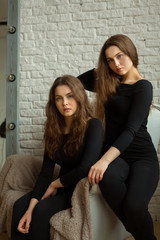 two beautiful young girl sisters twins with flowing hair in black clothes against a white brick wall background