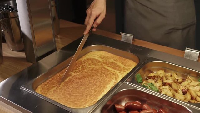 Close up shot of cook cutting big casserole in a food tray with a long knife. He slowly cuts it verticaly.