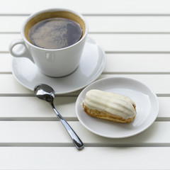 Fresh eclair with white chocolate and cup of espresso, black coffee on white wooden table, top view