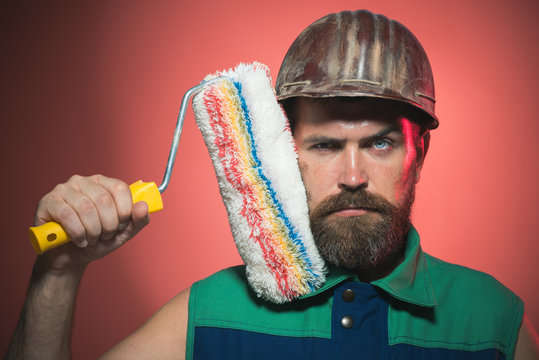 Labour and repairing concept. Bearded painter man, brutal caucasian hipster with beard and moustache wears helmet and holds paint roller near face. Man worker. Construction. Closeup portrait.