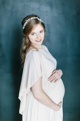 Young beautiful pregnant woman with a beautiful diadem in the hair poses standing on dark background. © sofiko14