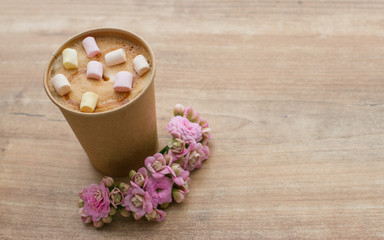 Coffee to go in paper cup on wooden background with pink flowers, place for text