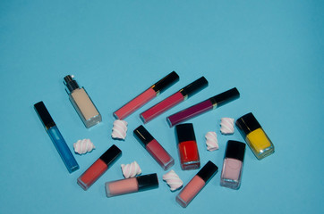 .Red, violet, pink and blue lipsticks. Сoncealer, red, pink and yellow nail polish. Make up beauty products on blue background near pink and white marshmallows. Top view, flatlay. Place for text.