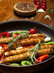Fried sausages with herbs, spices and vegetables in a pan. On wooden background.