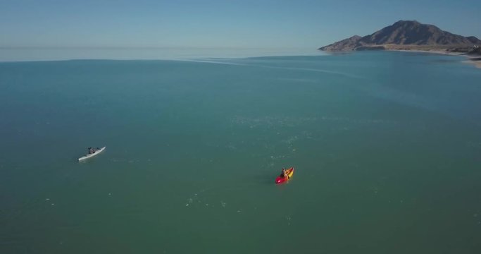 Aerial orbit around a young couple in kayaks talking and setting in the water off a sandy beach.