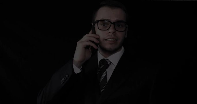 Handsome young businessman wearing glasses, talking on the phone in a suit, black background.