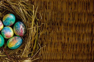 Many multi-colored Easter eggs lie in a nest of dry grass and twigs. Nest with eggs lie on the background of a table made of wicker willow twigs.
