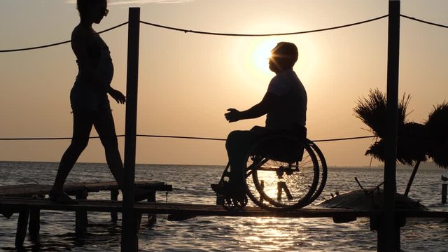 romantic date of female with male invalid on wheelchair on jetty near sea water against beautiful summer sunset